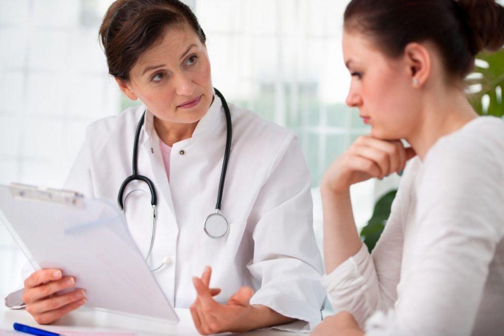 Female doctor holding clipboard speaking with female patient.