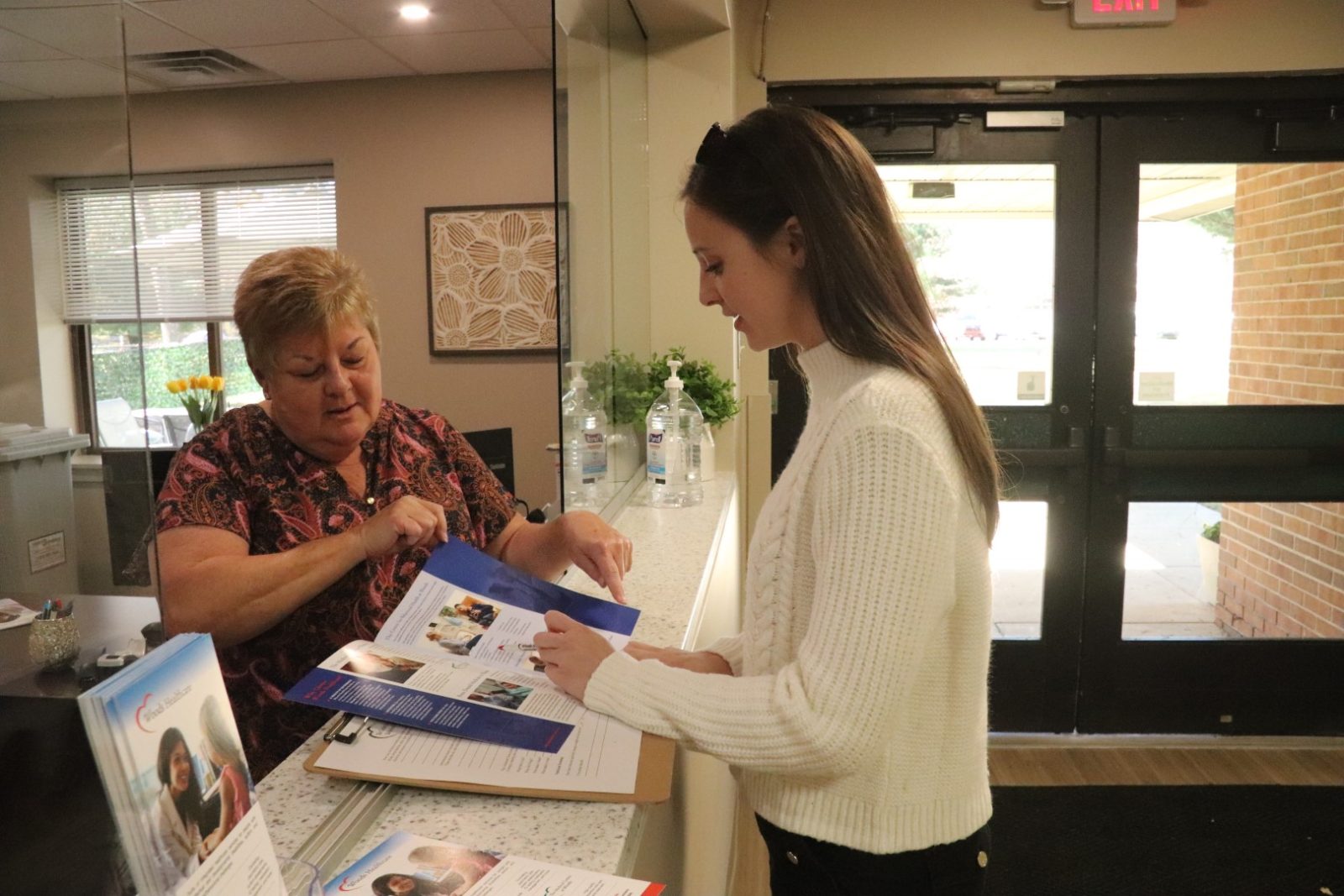 A female patient holding a pamphlet while speaking with receptionist at front desk.